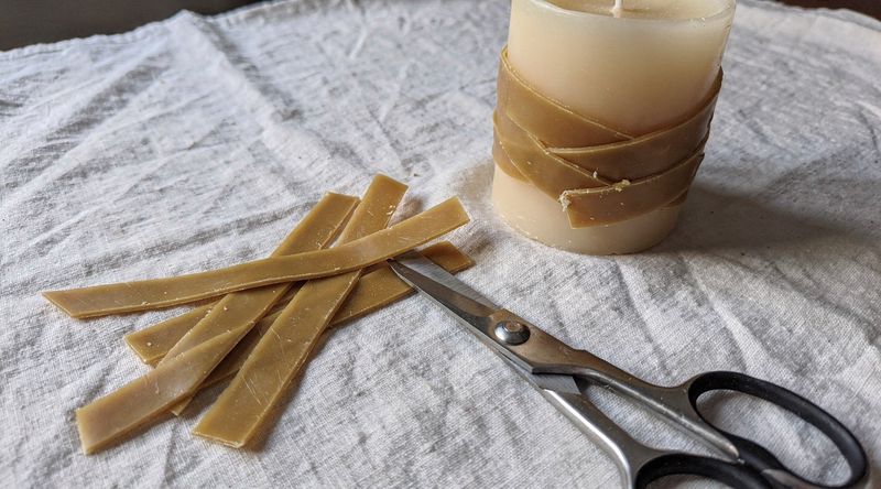 Hände schneiden ein geometrisches Muster aus einer goldenen Wachsplatte, daneben liegt eine halbfertige Kerze