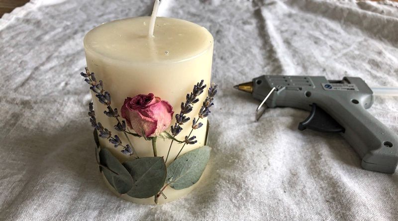 Nahaufnahme: Getrocknete Rose wird auf eine cremefarbene Kerze geklebt, umgeben von Lavendel und Eukalyptus